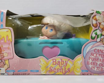 May include: A vintage "Baby Scents" doll in a blue tub, packaged in a pink box. The doll has blonde hair and blue eyes. The box features illustrations of children and candy. The text on the box reads "Candy Scented Poseable Baby Doll in Tub."
