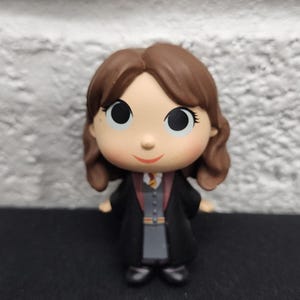 May include: A figurine of a character with brown hair, wearing a black robe with a gray vest and a red tie. The figure has large eyes and a small smile. The background is a white brick wall.