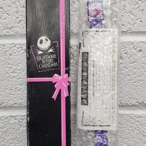 May include: A black box with a pink ribbon and the words "Nightmare Before Christmas" featuring Jack Skellington. Next to it is a purple item in bubble wrap. The item has a purple strap with a floral design.