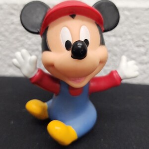 May include: A figurine of Mickey Mouse dressed as Mario, with a red cap and overalls. The figure has black ears, a black nose, and white gloves. The figure is sitting with its arms outstretched and yellow shoes.