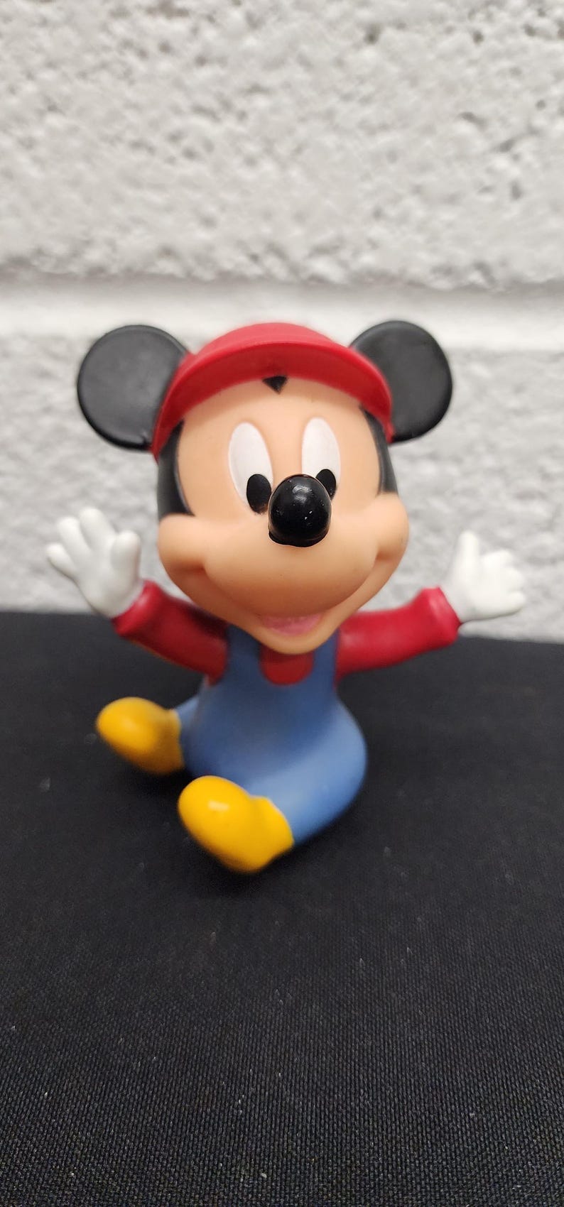May include: A figurine of Mickey Mouse dressed as Mario, with a red cap and overalls. The figure has black ears, a black nose, and white gloves. The figure is sitting with its arms outstretched and yellow shoes.