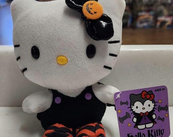 May include: A white plush Hello Kitty doll dressed in a black outfit with orange and black striped shoes. The doll has a black bow with an orange pumpkin accent. A purple tag with a Hello Kitty graphic is attached.