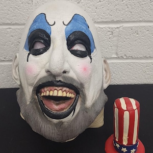 May include: A white clown mask with black and blue accents, a gray beard, and an open mouth. Next to it is a small red, white, and blue top hat with stars.