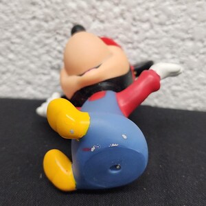 May include: A Mickey Mouse figurine in a relaxed pose, wearing his signature blue shorts, red shirt, yellow shoes, and black gloves. The figure has a red hat and is lying on a black surface.