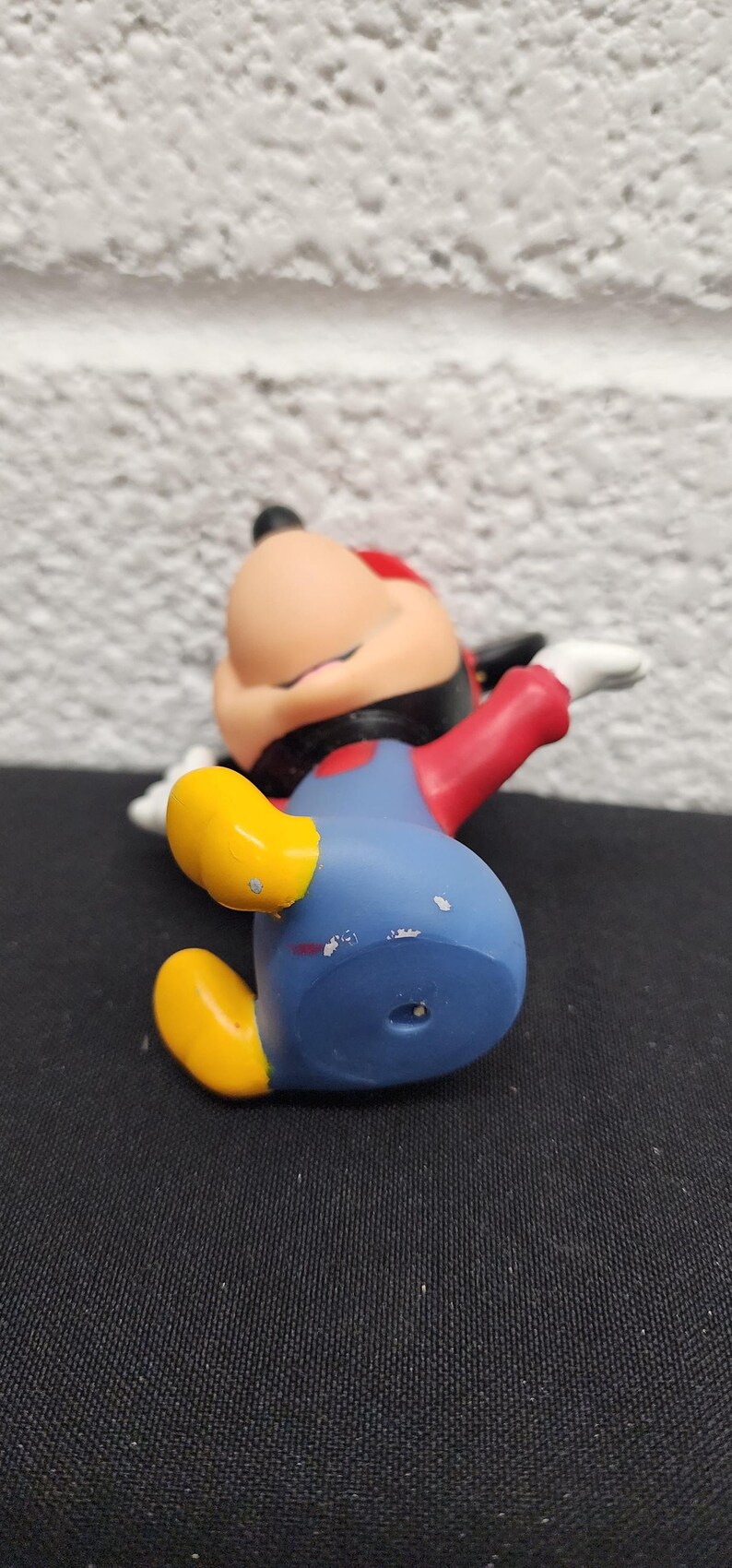 May include: A Mickey Mouse figurine in a relaxed pose, wearing his signature blue shorts, red shirt, yellow shoes, and black gloves. The figure has a red hat and is lying on a black surface.