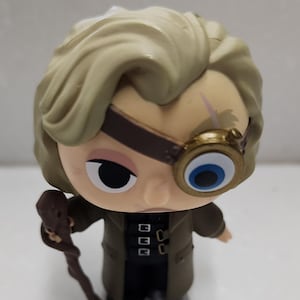 May include: A small plastic figurine of a character from the Harry Potter series. The figurine is wearing a brown coat and has a monocle over one eye. The figurine is holding a brown walking stick.