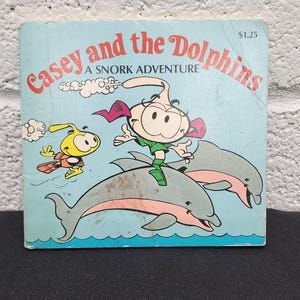 Puede incluir: Un libro infantil vintage titulado "Casey and the Dolphins: A Snork Adventure". La portada presenta ilustraciones de dibujos animados de un personaje montando delfines, con un personaje amarillo volando cerca. El libro tiene un fondo azul.