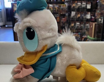 May include: A plush Donald Duck toy in a seated position. The toy features a white body, a blue sailor suit, a yellow beak and feet, and a blue sailor hat. A pink bow is visible. The toy has large blue eyes.