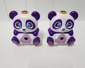 May include: Two white and purple panda toys with a heart-shaped opening on the front. The pandas have black eyes and a gold crown on top of their heads.