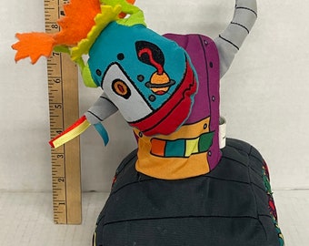 May include: A colorful plush robot toy with a black base and a teal head. The robot has orange, green, and red accents, with a gray arm raised. The robot's head features a unique design with a planet and a red swirl. A wooden ruler is next to the toy.