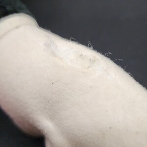 May include: Close-up of a plush toy's white arm with a black sleeve. The arm has a small tear with visible threads and a few dark specks. The background is a dark, solid color.