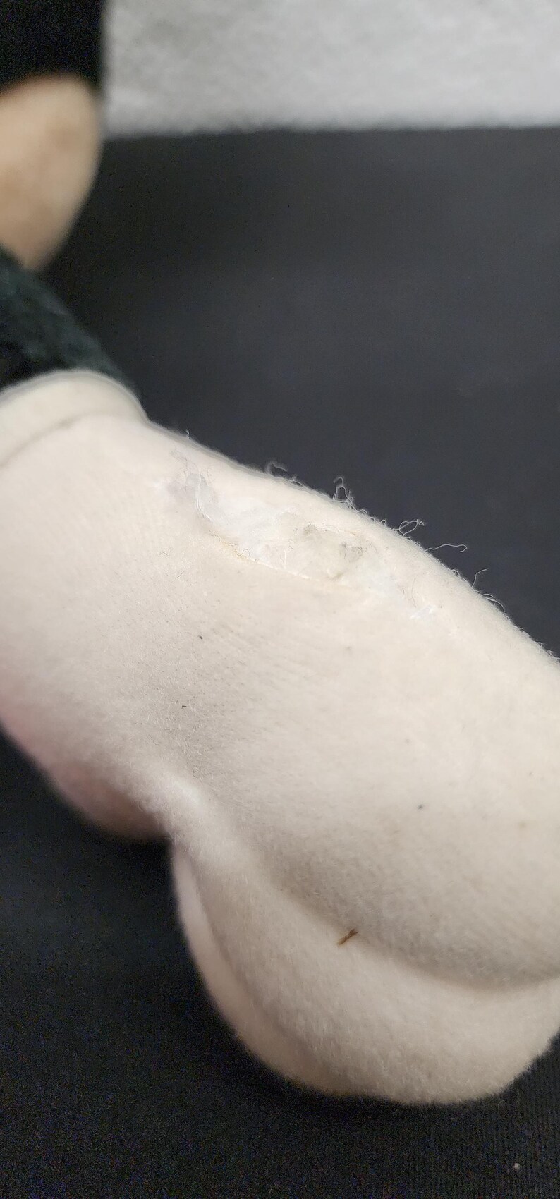 May include: Close-up of a plush toy's white arm with a black sleeve. The arm has a small tear with visible threads and a few dark specks. The background is a dark, solid color.