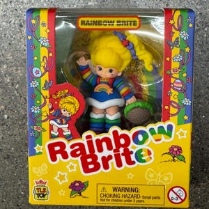 May include: A Rainbow Brite doll in its original packaging. The box is yellow with the words "Rainbow Brite" in large, colorful letters. The doll has yellow hair and a rainbow-colored outfit. The box also has a warning about choking hazards.