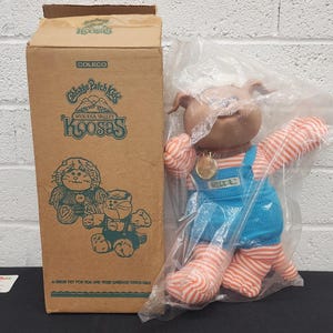 May include: A vintage Cabbage Patch Kids Koosas doll in a plastic bag next to its original box. The doll has a brown head, blue overalls, and striped arms and legs. The box features the Koosas logo and illustrations.
