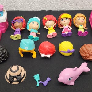 May include: A collection of colorful dollhouse accessories and figurines. Includes dolls with various hairstyles and outfits, a pink bed, a sink, and other decorative items. The accessories are in various colors, including pink, blue, yellow, and brown.