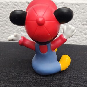 May include: A figurine of a cartoon character wearing a red hat, red shirt, blue overalls, and yellow shoes. The character has black ears and white gloves, with arms outstretched. The figurine is on a black surface.