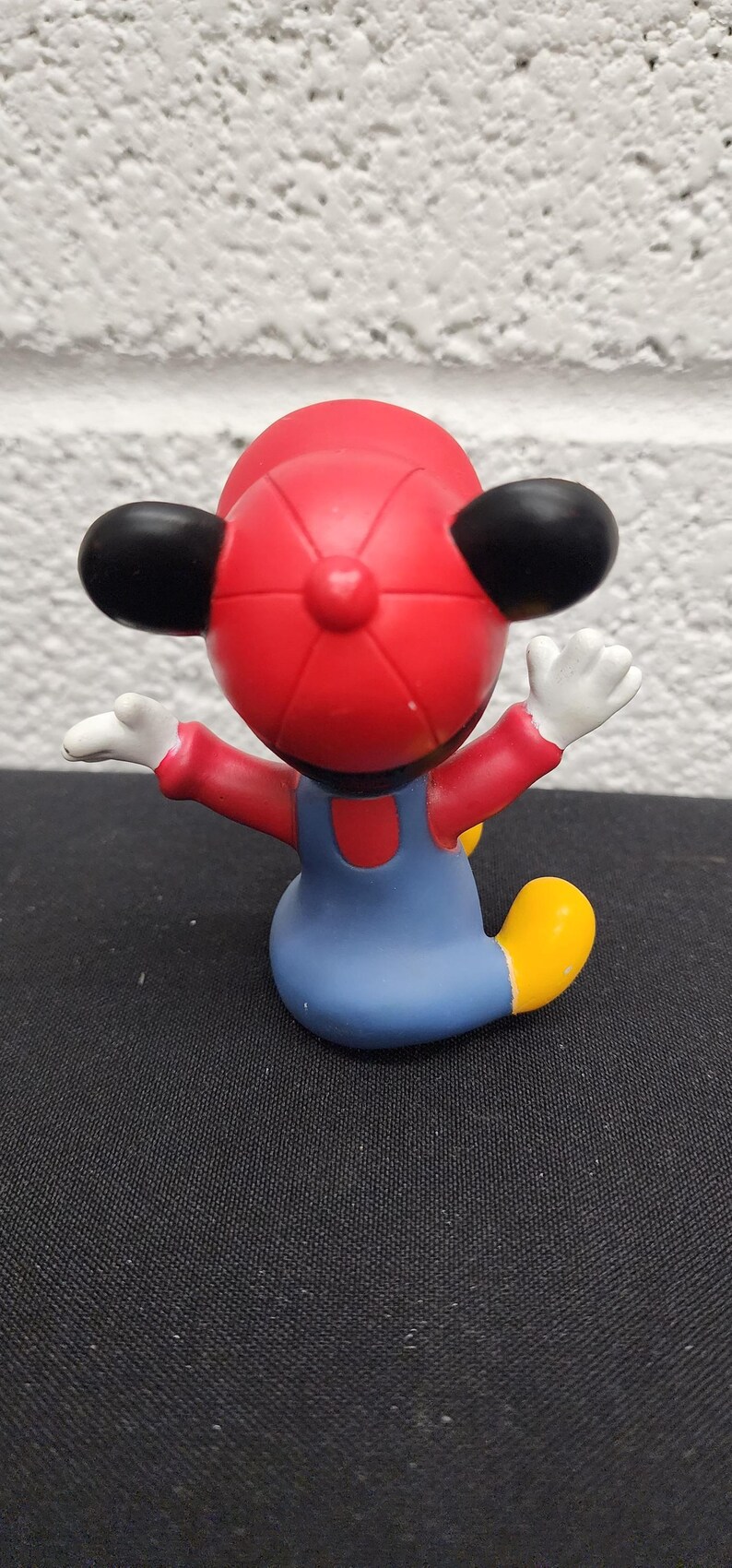 May include: A figurine of a cartoon character wearing a red hat, red shirt, blue overalls, and yellow shoes. The character has black ears and white gloves, with arms outstretched. The figurine is on a black surface.