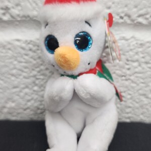 May include: A white plush snowman toy with a red Santa hat and a green and red scarf. The snowman has large blue glittery eyes and an orange beak. The snowman is sitting with its hands clasped.