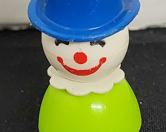 May include: A vintage green and white plastic toy clown with a blue top hat. The clown has a red nose and a painted smile.