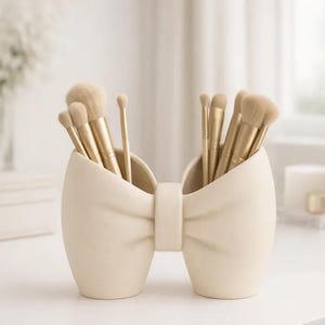 May include: Cream-colored bow-shaped makeup brush holder filled with various gold-handled brushes. The holder is made of ceramic and sits on a white surface. The brushes are arranged neatly, suggesting a focus on beauty and organization.