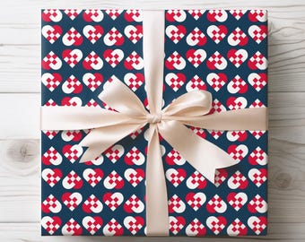 May include: A square gift box wrapped in dark blue paper featuring a repeating pattern of red and white woven heart shapes. The package is tied with a smooth, light beige satin ribbon that forms a centered bow across the top surface.