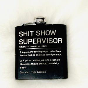 May include: Black stainless steel flask with a silver cap and hinge. The flask has the words "SHIT SHOW SUPERVISOR" in silver lettering, along with a definition. See also: Tina Gresian.