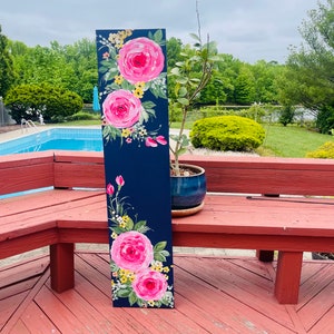 May include: A hand-painted wooden sign with a dark blue background and pink roses with yellow centres. The sign is leaning against a red wooden bench.