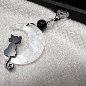 May include: A decorative phone charm featuring a black cat silhouette perched on a white crescent moon. The moon has a pearlescent finish. A black bead and clear crystal accents are attached to the charm.