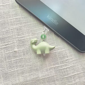 May include: A light green dinosaur-shaped bookmark attached to a Kindle e-reader. The bookmark has a small green bead and a clear bead. The e-reader is black and has the word "kindle" printed on it.