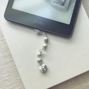 May include: A beaded Kindle charm featuring a small white elephant charm, gray and white beads, and clear crystal beads. The charm is attached to a black Kindle device, resting on a white book.