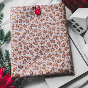 May include: A beige fabric book cover with a repeating pattern of gingerbread men and candy canes. The cover has a red button closure.