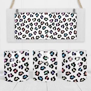 May include: Three white ceramic mugs featuring a colourful leopard print design. The mugs are arranged in a row on a white wooden surface.