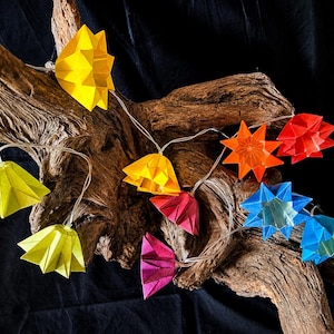 Könnte beinhalten: Eine Lichterkette mit dekorativen, geometrischen, gefalteten Lampenschirmen in Gelb, Orange, Rot, Pink und Blau. Die Lichter sind durch ein dünnes, transparentes Stromkabel miteinander verbunden und über ein Stück Treibholz drapiert.