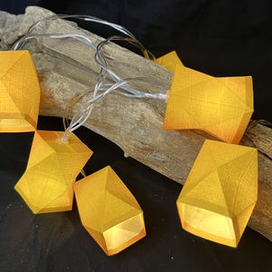 Könnte beinhalten: Eine LED-Lichterkette mit geometrischen, facettierten, goldgelben Lampenschirmen. Die Lichter sind durch einen dünnen, transparenten Draht verbunden und über ein Stück verwittertes, hellbraunes Treibholz drapiert, was einen warmen Schein erzeugt.