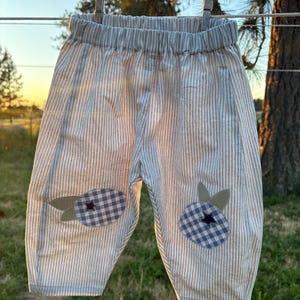 May include: Light blue and white pinstriped trousers featuring an elastic waistband and two decorative knee patches. Each patch is shaped like a blueberry with a blue and white gingham pattern, accented by two small green leaves and a dark star.