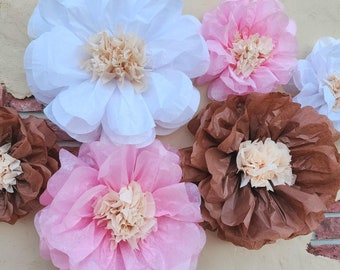 May include: A collection of six tissue paper flowers in various colors, including white, pink, and brown. The flowers are arranged on a wall, creating a decorative display.