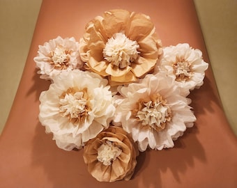 May include: A bouquet of six tissue paper flowers in shades of brown and white. The flowers are arranged in a circular pattern on a peach-colored surface.