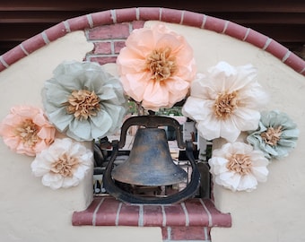 May include: A rustic metal bell with a black finish hangs from a wrought iron frame. The bell is surrounded by a variety of pastel-colored paper flowers, including peach, light blue, and cream.