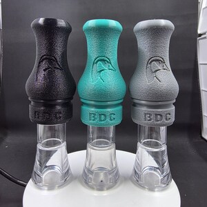May include: Three duck calls in black, teal, and gray. Each call features a textured grip, a duck logo, and the letters "BDG". The calls have clear acrylic barrels.