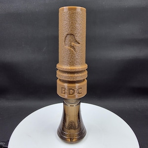 May include: A brown duck call with a textured surface and a duck head logo. The call has a cylindrical top, a middle section with the letters "BDC", and a flared bottom. The call is made of plastic and metal.