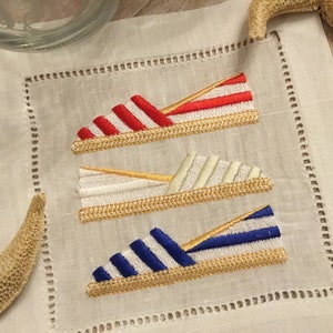 May include: Three embroidered linen napkins with a nautical theme. Each napkin features a different colored striped espadrille shoe with a gold rope detail.