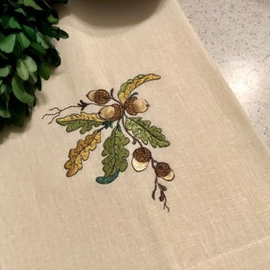 May include: A beige linen tea towel with an embroidered design of a branch with green and gold leaves and brown acorns.