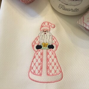 May include: Pink embroidered kitchen towel with a Santa Claus design holding a gift. The towel has a textured surface and the Santa is wearing a pink and white suit with a white beard and a red hat.