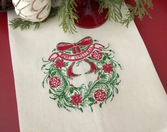 May include: White linen kitchen towel with a red and green embroidered Christmas wreath design. The wreath has a red bow and a banner that reads "Merry Christmas".