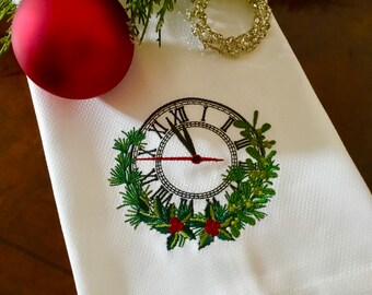 May include: White kitchen towel with an embroidered design of a clock face surrounded by holly and greenery. The clock face is black with Roman numerals and the hands are pointing to midnight.