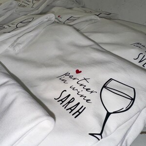 May include: A stack of white t-shirts with a black and red design that says "partner in wine SARAH" with a small red heart and a drawing of a wine glass.