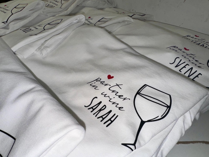 May include: A stack of white t-shirts with a black and red design that says "partner in wine SARAH" with a small red heart and a drawing of a wine glass.