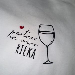 May include: White t-shirt with black text that reads "partner in wine RIEKA" and a red heart above the text. A black outline of a wine glass is to the right of the text.