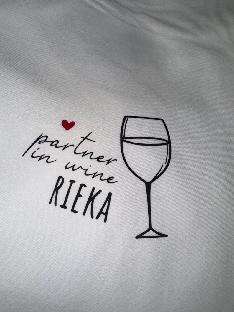 May include: White t-shirt with black text that reads "partner in wine RIEKA" and a red heart above the text. A black outline of a wine glass is to the right of the text.