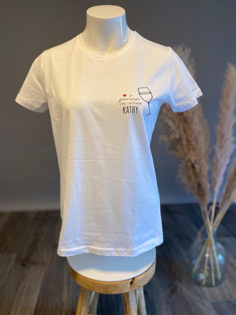 May include: White t-shirt with a black graphic of a wine glass and the text "partner in wine KATHY".